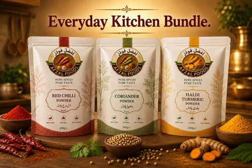 Three stand-up pouches of AFZAL FOODS spices arranged on a wooden surface with kitchen ingredients around them: Red Chilli Powder, Coriander Powder, and Haldi Turmeric Powder. The pouches are shown in a warm kitchen-style setting with spices and herbs, and the text 'Everyday Kitchen Bundle.